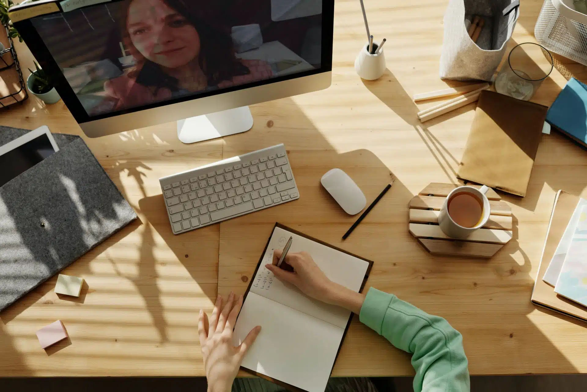 Aerial shot of people taking notes while watching a computer screen on a desk, stationery and a coffee cup are on the desk